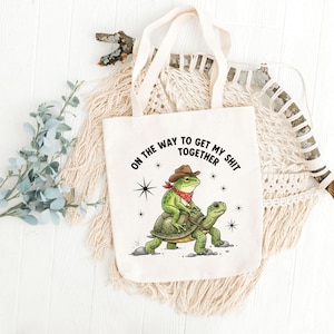 May include: Off-white tote bag with a whimsical design. A green frog wearing a cowboy hat and red bandana rides a green turtle. The text "ON THE WAY TO GET MY SHIT TOGETHER" is printed above the frog and turtle.