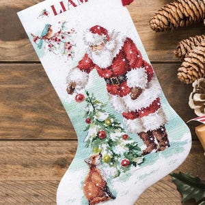 May include: A white Christmas stocking featuring a cross-stitched design of Santa Claus decorating a tree with a fox nearby and a blue bird on a branch. The name LIAM is embroidered in red at the top. A small red tassel is attached to the upper corner.