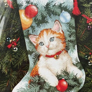 Könnte beinhalten: Ein Weihnachtsstrumpf mit einem Kreuzstichmotiv eines orange-weißen Kätzchens, das ein rotes Halsband mit einer Glocke trägt. Das Kätzchen ist von grünen Tannenzweigen und bunten Ornamenten umgeben. Der Name Bethany ist oben in Braun eingestickt.