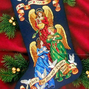 May include: A navy blue Christmas stocking with embroidered angels and the name "Christina" at the top. The angels are depicted in red, green, and blue gowns with golden wings. The phrase "Hark the Herald Angels Sing" is also embroidered on the stocking.