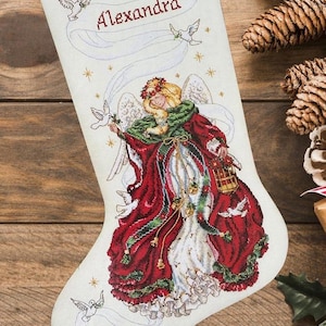 Könnte beinhalten: Ein weißer Weihnachtsstrumpf mit einem im Kreuzstich gestickten Engel in einem rot-grünen Gewand, der eine Laterne hält. Das Design zeigt weiße Tauben, goldene Sterne und ein Banner mit dem Namen Alexandra, der in rotem Garn eingestickt ist.