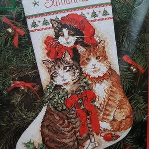 May include: A white Christmas stocking featuring a cross-stitch design of three cats. The top border has red and green stripes with the name Samantha embroidered in red. The cats are wearing a red Santa hat, a green wreath, and a red bow.