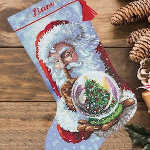 Puede incluir: Un calcetín navideño con un diseño de punto de cruz de Santa Claus sosteniendo una bola de nieve con un árbol de Navidad dentro. El nombre Liam está bordado en rojo en la parte superior. El calcetín tiene un fondo azul claro y una borla roja.