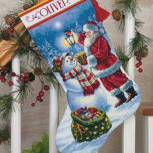 May include: A Christmas stocking featuring a winter scene with Santa Claus, a snowman, and a bag of gifts. The stocking is blue with a red border and the name "OLIVER" at the top. The scene is set in a snowy landscape.