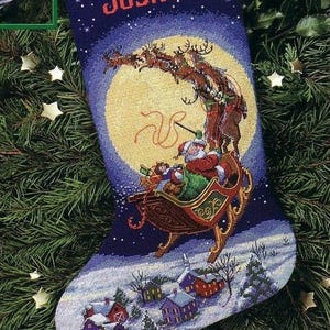 以下が含まれることがあります： サンタクロースがトナカイに引かれるそりに乗っている夜のシーンが描かれたクリスマスストッキング。上部には赤い文字で「Joshua」と書かれています。ストッキングの下部には雪の降る村のシーンがあり、背景には満月があります。