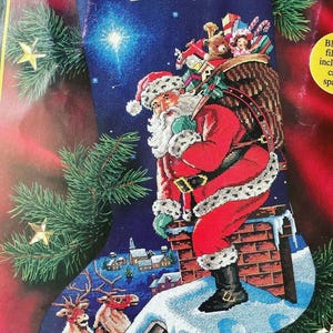 Peut inclure: Une chaussette de Noël bleu foncé avec un motif au point de croix représentant le Père Noël sur une cheminée en briques avec un sac de jouets. Le nom Duncan est inscrit en haut. Deux rennes sont sur un toit enneigé avec un village au loin.