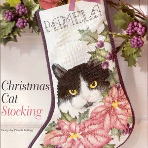 May include: A Christmas stocking featuring a black and white cat face surrounded by pink poinsettias and green holly leaves. The word "PAMELA" is stitched at the top. The text "Christmas Cat Stocking" is also visible.