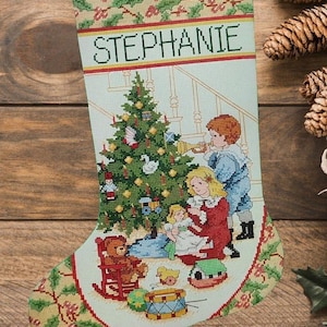 Könnte beinhalten: Ein Weihnachtsstrumpf mit einer Kreuzstichszene eines geschmückten Baumes, eines Kindes mit Trompete und eines Kindes mit Puppe. Der Name STEPHANIE ist in schwarzen Buchstaben auf hellblauem Grund mit Stechpalmen-Bordüren eingestickt.