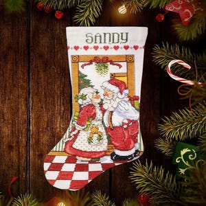 May include: A white Christmas stocking with the name "SANDY" embroidered at the top. The stocking features a cross-stitch design of Santa Claus and Mrs. Claus. The background is a wooden surface with Christmas decorations.