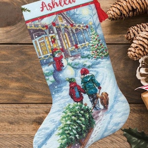 Puede incluir: Un calcetín navideño con una escena invernal en punto de cruz que muestra una casa, un muñeco de nieve, dos figuras, un perro y un pino sobre un trineo. El nombre Ashlea está bordado en rojo en la parte superior. Incluye un lazo rojo con borla.