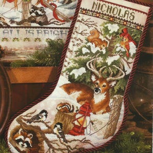 May include: A Christmas stocking with a cross-stitch design featuring a deer, squirrel, birds, and a red lantern in a snowy forest scene. The name "NICHOLAS" is stitched at the top, with a red rope trim.
