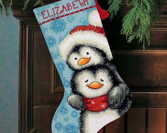 May include: A Christmas stocking with a light blue background and snowflake pattern. The stocking features two penguins wearing Santa hats and red scarves. The name "ELIZABETH" is embroidered in red at the top. A red loop is attached for hanging.