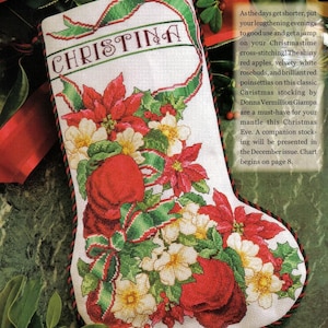 Pode incluir: Uma meia de Natal com o nome "CHRISTINA" bordado em verde. A meia é decorada com poinsétias vermelhas, botões de rosa brancos e folhas de azevinho verdes. Uma fita vermelha e verde emoldura a parte superior.