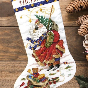 Puede incluir: Un calcetín navideño blanco con un diseño de punto de cruz de Santa Claus cargando un árbol verde y un saco de juguetes. El borde superior es azul oscuro con estrellas amarillas y el nombre NICHOLAS impreso en color blanco.