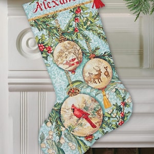 May include: Christmas stocking with the name "Alexander" embroidered in red. The stocking features three round ornaments with winter scenes, including a red cardinal, deer, and a farmhouse. The background is a light blue with green foliage.