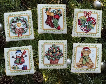 May include: Six square fabric ornaments featuring cross-stitch designs of a Christmas wreath, two stockings, a heart, a snowman, a potted tree, and a teddy bear. Each white square is bordered by a gold frame and green leaf pattern.