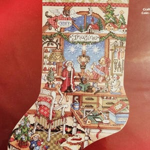 May include: Cross-stitch Christmas stocking featuring a festive workshop scene. The design includes Santa Claus, a teddy bear holding a sign that says "Dream", and the word "JOYCE" at the top. The stocking is filled with holiday cheer.