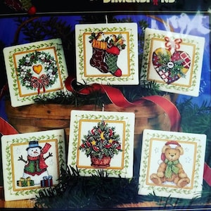 May include: A collection of six square cross-stitch ornaments with Christmas themes. Designs include a wreath, stockings, a heart, a snowman, a Christmas tree, and a teddy bear, all framed with a gold border and green garland.