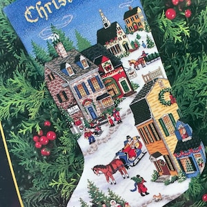 Puede incluir: Un calcetín navideño con un diseño de punto de cruz que representa una escena de un pueblo invernal. La parte superior del calcetín es azul con el nombre "Christopher" bordado en dorado. La escena incluye casas, personas y un trineo tirado por caballos.