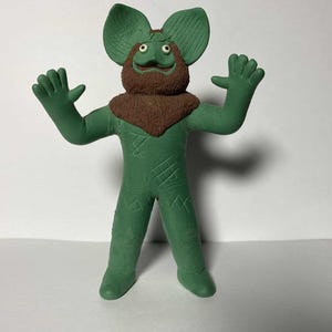 Puede incluir: Una figura verde de una criatura con orejas grandes, barba marrón y brazos extendidos. La figura tiene una superficie texturizada y está de pie. Los ojos de la criatura son pequeños y redondos.