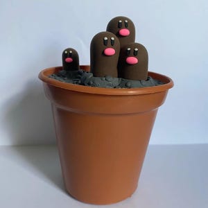 Può includere: Un vaso di terracotta marrone con tre figure di terracotta marrone con guance rosa e occhi neri. Le figure sono parzialmente sommerse nel terreno di argilla grigia.