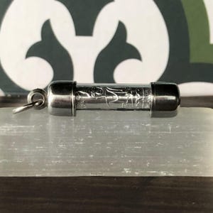 Puede incluir: Un vial de vidrio transparente con tapas de metal plateado y un lazo. El vial contiene un diseño grabado. El vial descansa sobre una piedra blanca rectangular. El fondo es un diseño de patrón verde y blanco.