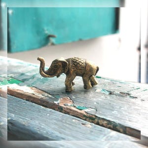 May include: A small, antique-style brass elephant figurine. The elephant has detailed features, including a raised trunk and textured skin. It stands on a weathered, blue-painted wooden surface, with a turquoise background. The figurine is a decorative item.