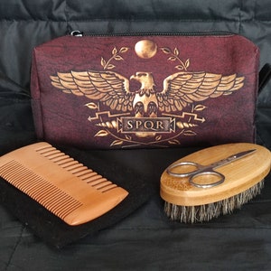 Puede incluir: Una bolsa de aseo roja con un diseño de águila dorada y la palabra "SPQR" impresa en ella. La bolsa está junto a un peine de madera, un cepillo de madera y un pequeño par de tijeras.