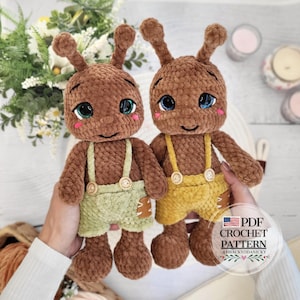 May include: Two brown plush crochet dolls with antennae, large blue and green eyes, and pink cheeks. One doll wears light green overalls with wooden buttons, and the other wears yellow overalls with wooden buttons. Text reads: PDF CROCHET PATTERN.
