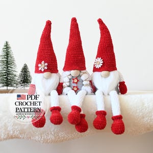 Op de afbeelding: Drie gehaakte gnomen met rode mutsen, witte baarden en grijze lichamen. De gnomen zitten op een wit, pluche oppervlak. De gnome links heeft een sneeuwvlok op zijn muts, de gnome in het midden heeft een rode knoop en een ster op zijn lichaam, en de gnome rechts heeft een witte bloem op zijn muts. De gnomen dragen witte gehaakte wanten.