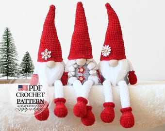 Peut inclure: Trois gnomes en crochet avec des chapeaux rouges, des barbes blanches et des corps gris. Les gnomes sont assis sur une surface blanche et moelleuse. Le gnome de gauche a un flocon de neige sur son chapeau, le gnome du milieu a un bouton rouge et une étoile sur son corps, et le gnome de droite a une fleur blanche sur son chapeau. Les gnomes portent des mitaines en crochet blanches.