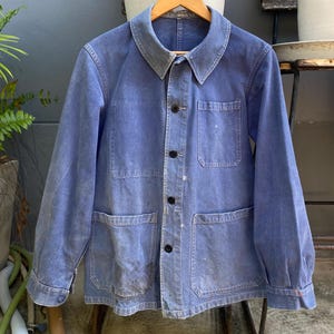 Peut inclure: Veste en jean bleu délavé avec un col classique et quatre poches avant. La veste se ferme avec des boutons et a des manches longues. Le denim présente des signes d'usure, avec des taches et un aspect usé, suggérant qu'elle est vintage.