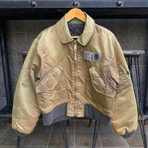 May include: A vintage-style, olive green bomber jacket with a zip fastening and a patch on the left chest. The jacket has a ribbed knit collar, cuffs, and waistband.