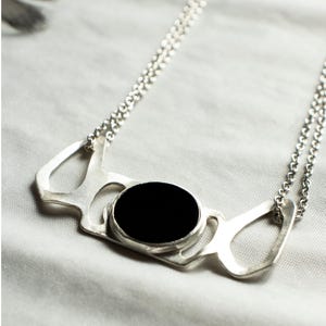 May include: A silver necklace with a large, oval black stone pendant. The pendant has an abstract, openwork design. The necklace chain is delicate and the background is a soft, white fabric.