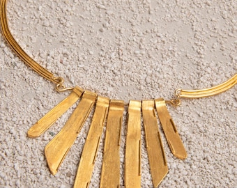 May include: A gold-colored necklace with a series of vertical, rectangular pendants fanning out from a curved collar. The pendants have a textured surface and are arranged in a symmetrical pattern. The necklace is displayed on a light-colored, textured surface.