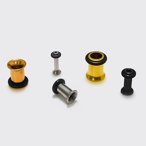 May include: Five ear gauges in different designs and colours. The gauges include gold, silver, and black options. Some have a flared design, while others are cylindrical. The gauges are displayed on a white surface.
