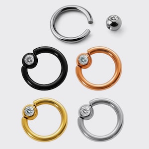 May include: An assortment of body jewelry, including a silver captive bead ring, a silver ball, and four captive bead rings in black, rose gold, gold, and silver. Each ring features a clear, round gemstone.