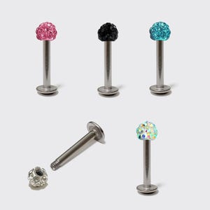Può includere: Una collezione di gioielli per piercing alle labbra con top colorati tempestati di cristalli. I gioielli presentano un perno e una base argentati, con top rosa, neri, blu e iridescenti. Un pezzo è smontato.