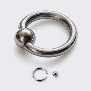 Puede incluir: Un gran anillo de cuentas cautivas plateado brillante con una cuenta a juego y un anillo y una cuenta más pequeños. La joyería se exhibe sobre un fondo blanco. El anillo es un piercing corporal.