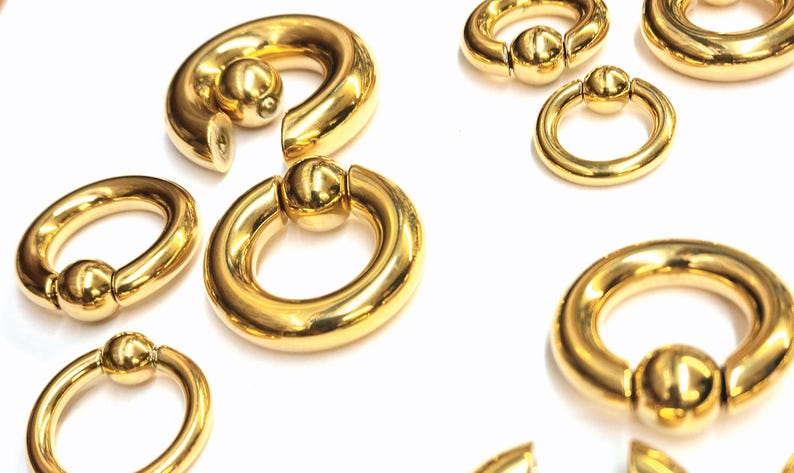 May include: A set of gold-colored metal body jewelry, including circular rings with a ball closure. The rings are of different sizes and are arranged on a white surface.