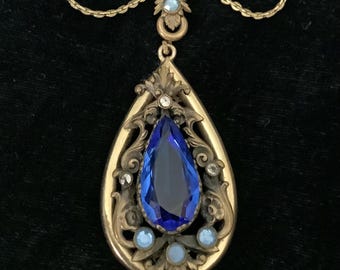 May include: A vintage gold-toned necklace with a large, teardrop-shaped pendant. The pendant features a faceted, deep blue gemstone, surrounded by ornate filigree and smaller, light blue stones. The chain is delicate and has a clasp.