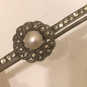 Op de afbeelding: Vintage zilverkleurige broche met een centrale parel en marcasiet accenten. De broche heeft een geweven ontwerp rond de parel en is gezet op een staaf met extra marcasiet stenen. Een klassiek sieraad.