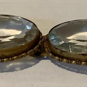 Pode incluir: Uma fivela dourada antiga com duas grandes pedras ovais, facetadas e transparentes. As pedras s&atilde;o fixadas numa moldura decorativa com contas. A fivela tem uma est&eacute;tica vintage.