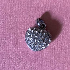Può includere: Un ciondolo a forma di cuore argentato incastonato con numerosi cristalli trasparenti. Il ciondolo ha un piccolo anello nella parte superiore per il fissaggio a una catena. Lo sfondo è rosa.