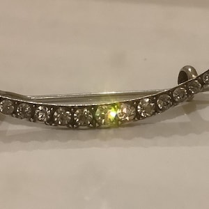 May include: A crescent-shaped brooch with a silver-tone finish, adorned with numerous clear, round gemstones. The brooch curves gracefully, with the gemstones set along its length. The design is elegant and suitable for formal wear.