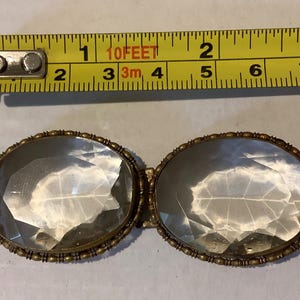 Pode incluir: Um pingente antigo de vidro transparente em forma oval com uma moldura de metal dourado. O pingente tem aproximadamente 5 cm de comprimento. Uma fita m&eacute;trica amarela est&aacute; no fundo.