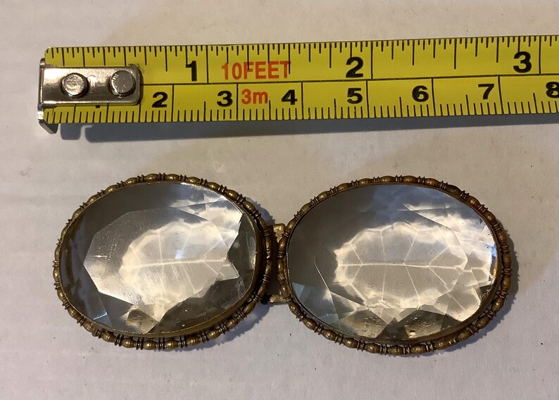 Pode incluir: Um pingente antigo de vidro transparente em forma oval com uma moldura de metal dourado. O pingente tem aproximadamente 5 cm de comprimento. Uma fita m&eacute;trica amarela est&aacute; no fundo.