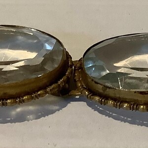 Pode incluir: Uma fivela dourada antiga com duas grandes pedras ovais, facetadas e transparentes. As pedras est&atilde;o emolduradas em uma moldura decorativa com contas. A fivela tem uma est&eacute;tica vintage.