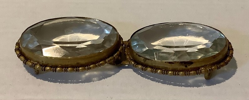 Pode incluir: Uma fivela dourada antiga com duas grandes pedras ovais, facetadas e transparentes. As pedras est&atilde;o emolduradas em uma moldura decorativa com contas. A fivela tem uma est&eacute;tica vintage.