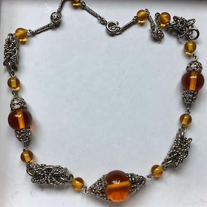 Puede incluir: Un collar vintage con cuentas de vidrio color ámbar y detalles metálicos plateados. El collar presenta una mezcla de cuentas redondas y oblongas, con cuentas decorativas más grandes. El trabajo en metal incluye filigranas y eslabones de cadena.